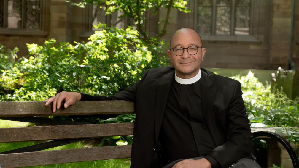 The Rev. Phillip A. Jackson, Rector sits on a wooden bench in the Trinity Churchyard. He has closely cropped hair, round, horn-rimmed glasses and is wearing a clerical collar under a black jacket.
