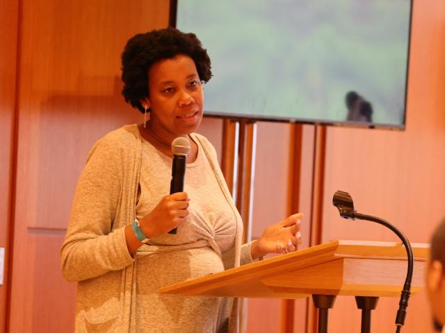 Rev. Jen Bailey speaks at a podium with a microphone in hand. She wears a light grey cardigan and dress with twist detail at her waist. She is a Black woman with a small afro.