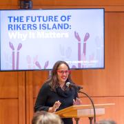 Susan Shah stands at a podium in front of digital signage that reads "The Future of Rikers Island: Why It Matters."
