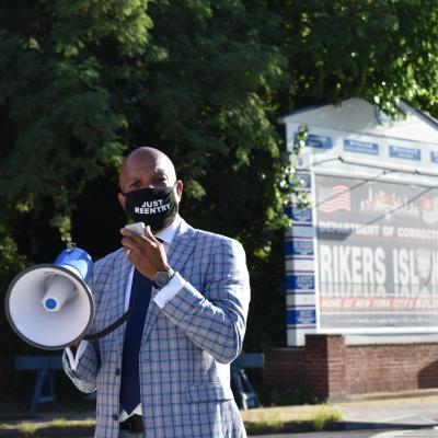 A masked man speaks into a megaphone while standing in front of a sign that says "Rikers Island."