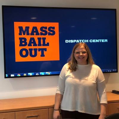 A young woman stands in front of a screen, which reads "Mass Bail Out Dispatch Center."
